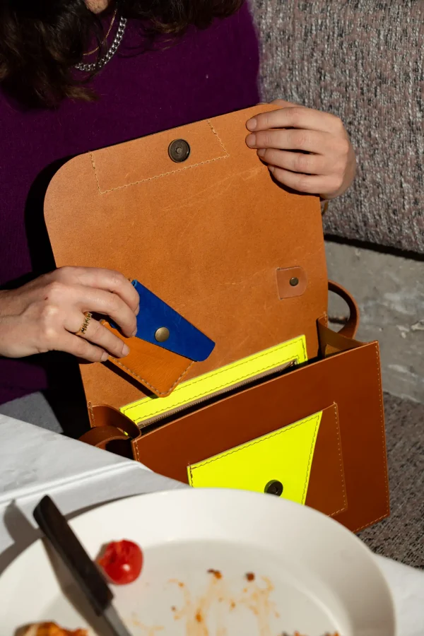 Vrouw steekt een cognac Halle B. portefeuille met kobaltblauw in een cognac Sofie L. handtas met fluogeel.
