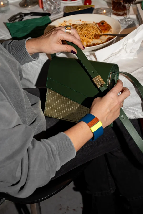 Vrouw heeft de groene Reese W. handtas met goud op haar schoot en draagt een kobaltblauwe The Combo armband met fluogeel en roest.