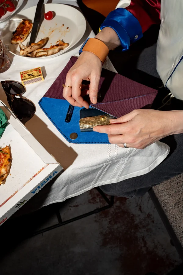Vrouw steekt de blauwe Halle B. portefeuille met goud in de paars gestreepte Billie E. enveloppetas en draagt een The Combo armband in naturel.