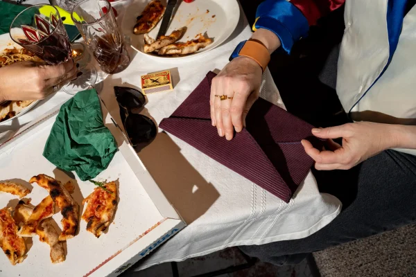 Vrouw heeft de paars gestreepte Billie E. enveloppetas in de hand en draagt een The Combo armband in naturel. Op de tafel ligt een Halle B. portefeuille in petrol en fluogeel.