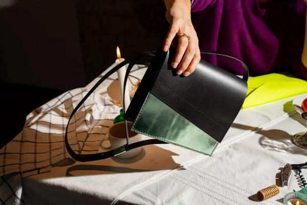 Vrouw neemt de zwarte Reese W. handtas met groen métallic front van de tafel. Er ligt ook een fluogele Billie E. enveloppetas op tafel.