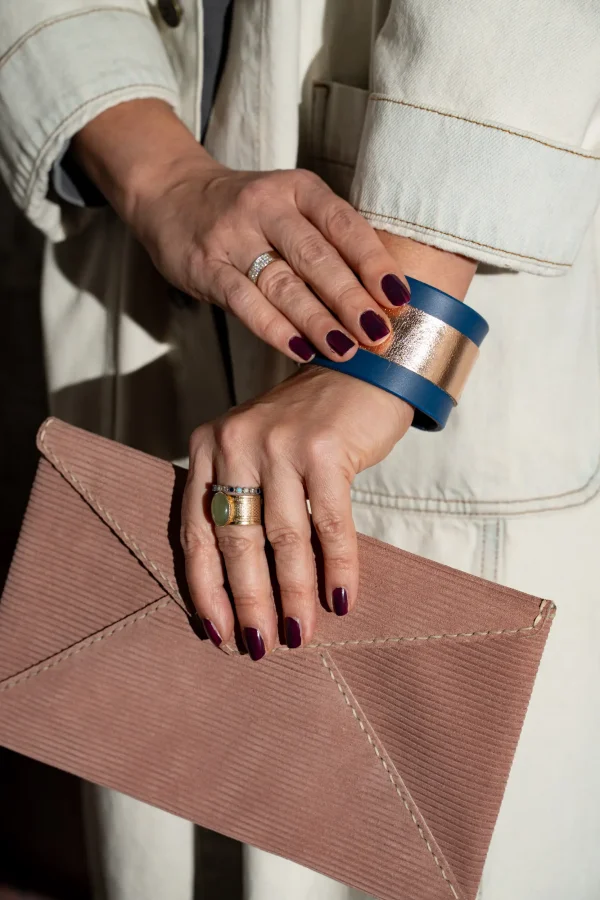 Vrouw heeft vuilroze gestreepte Billie E. enveloppetas in de hand en pruts ondertussen aan haar kobaltblauwe The Bold armband met rosé métallic contrast.
