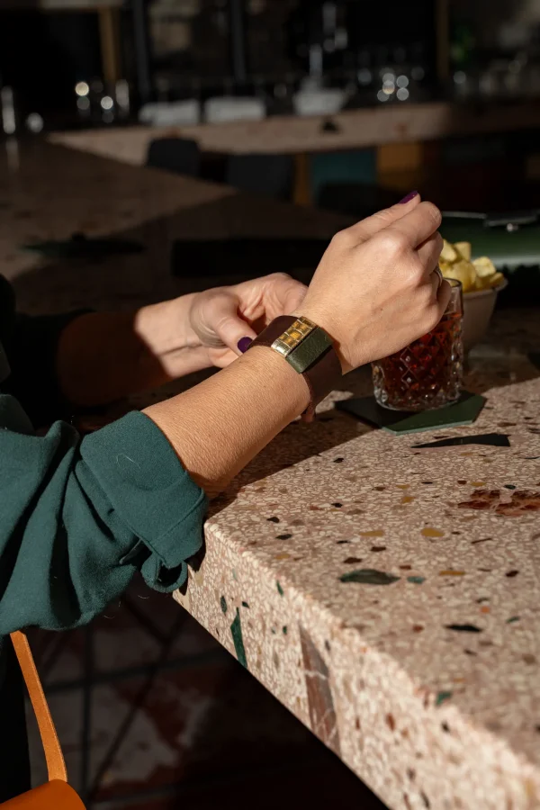 Vrouw doet haar bruine The Combo armband met goud en groen open. Haar glas staat op een groene coaster.