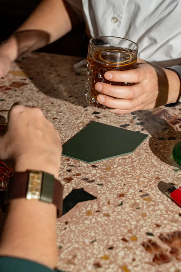 Twee vrouwen heffen het glas die op groene coasters staan. De ene vrouw draagt een The Combo armband, bruin met goud en groen.