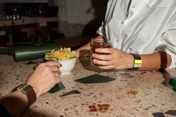 Twee vrouwen heffen het glas die op groene coasters staan. Ze dragen elke een The Combo armband, bruin met goud en groen en blauw met fluogeel en pistache. De groene Penélope C. handtas ligt op tafel.