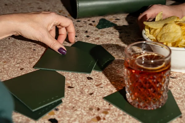 Groene coasters in asymmetrische vorm liggen op een tafel. Op eentje staat een glas. In de verte ligt de groene Penélope C. handtas.