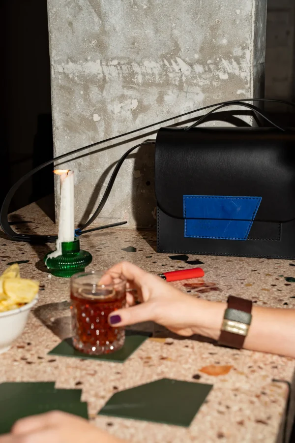 De zwarte Sofie L. met kobaltblauw contrast staat op een tafel terwijl iemand een glas vastheeft. Het glas staat op een groene coaster en de vrouw draagt een bruine The Combo armband met groen en goud.